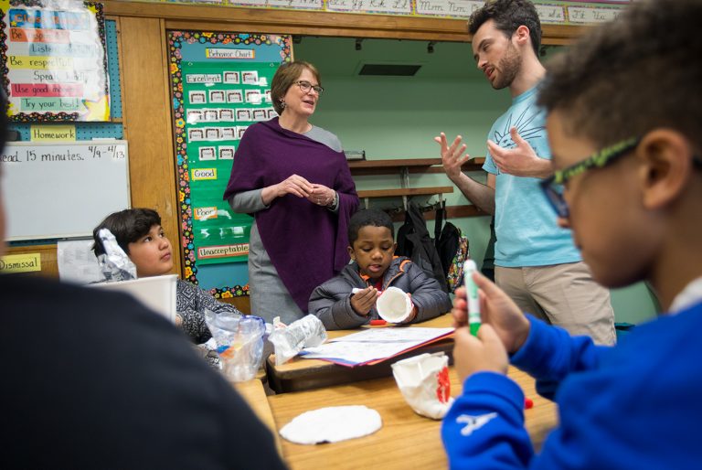 Christina H. Paxson and a man are engaged in conversation while three children work at school desks in front of them.