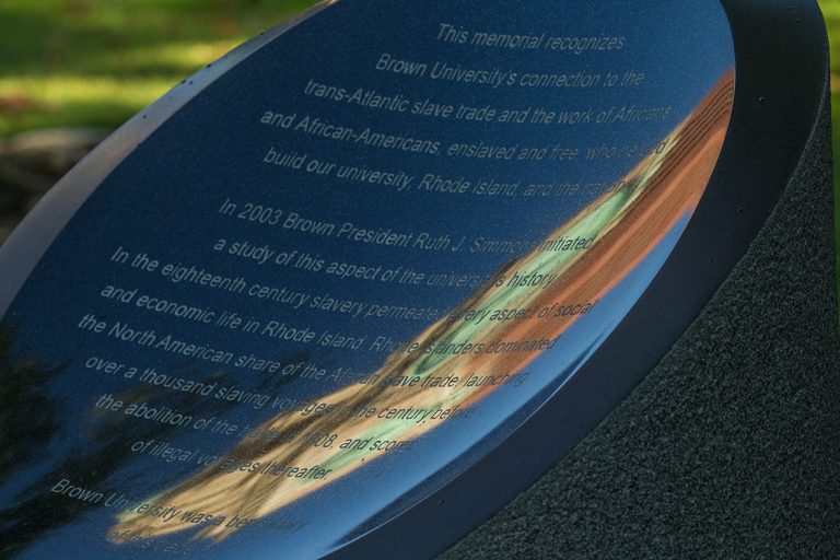 A grey stone plinth with engraved text that reads, "This memorial recognizes Brown University’s connection to the trans-Atlantic slave trade and the work of Africans and African-Americans enslaved and free who helped build our university, Rhode Island, and the nation. In 2003 Brown President Ruth J. Simmons initiated a study of this aspect of the university’s history. In the eighteenth century slavery permeated every aspect of social and economic life in Rhode Island. Rhode Islanders dominated the North American share of the African slave trade launching over a thousand slaving voyages in the century before the abolition of the trade in 1808 and scores of illegal voyages thereafter. Brown University was a beneficiary of this trade.”